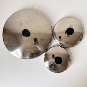 May include: Three stainless steel pot lids of varying sizes. Each lid is circular with a black, round handle in the center. The lids have a reflective surface, and the image reflects the person taking the photo.