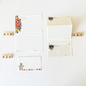 May include: A collection of vintage recipe cards. Some cards feature floral designs, a gingerbread man, and a bowl with a spoon. The cards have lined spaces for writing recipes. Wooden letter tiles spell out "A 19", "A 20", and "A 21".