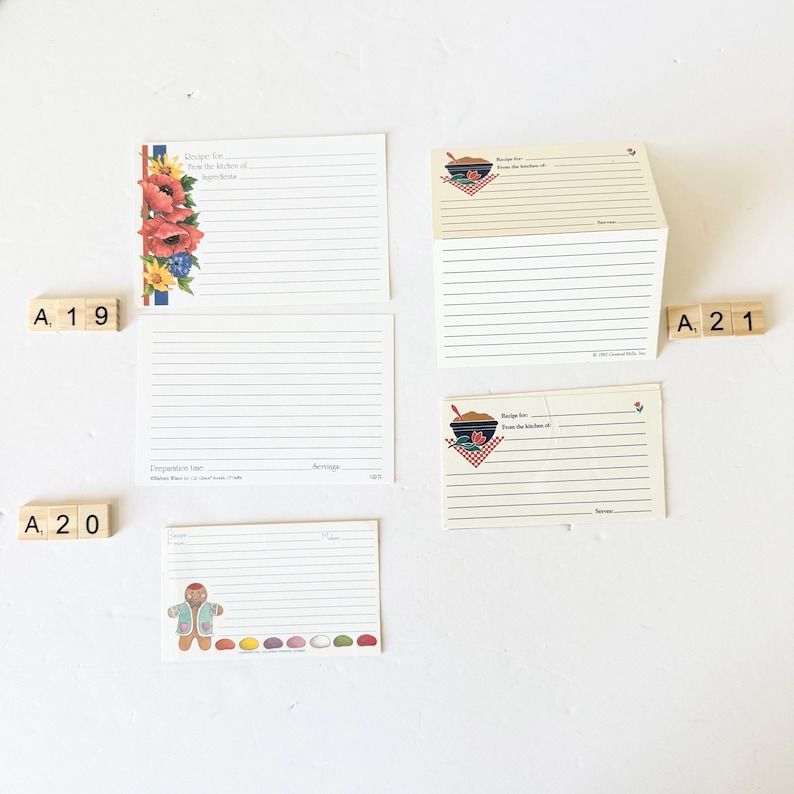 May include: A collection of vintage recipe cards. Some cards feature floral designs, a gingerbread man, and a bowl with a spoon. The cards have lined spaces for writing recipes. Wooden letter tiles spell out "A 19", "A 20", and "A 21".