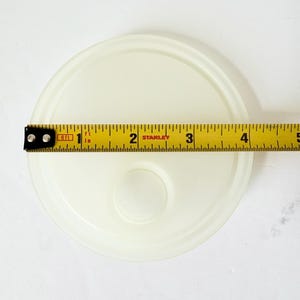 May include: A round, translucent off-white plastic lid featuring a raised circular center detail. A yellow Stanley measuring tape is positioned across the lid, indicating a diameter of approximately 4.5 inches for the circular object.