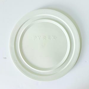 May include: A white Pyrex lid with a circular design. The lid features the embossed brand name "PYREX" in the center, with concentric rings and a raised edge. The words "CHINA" and "MADE IN USA" are also visible.