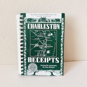 Może przedstawiać: Książka kucharska w oprawie spiralnej zatytułowana "Charleston Receipts" z zieloną okładką. Okładka zawiera mapę Charleston w Południowej Karolinie i tekst "Serving The Lowcountry For Over 60 Years."