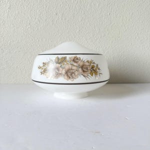 May include: White glass lampshade with a floral design in shades of brown, tan, and green. The lampshade has a rounded shape with a black trim and a small base. The floral pattern is centered around the lampshade.