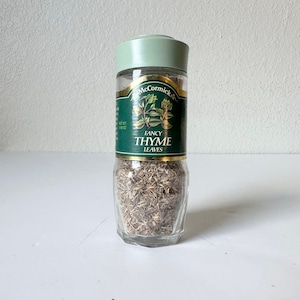 Może przedstawiać: Przezroczysty szklany słoik z przyprawami z zieloną pokrywką i etykietą. Na etykiecie napis "McCormick Fancy Thyme Leaves" z ilustracjami gałązek tymianku. Słoik jest wypełniony suszonymi liśćmi tymianku.