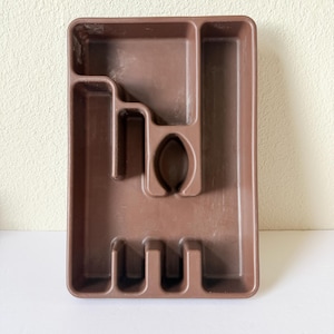 Puede incluir: Una bandeja de cubiertos de plástico marrón con múltiples compartimentos para organizar la cubertería. La bandeja tiene forma rectangular con varias ranuras y secciones diseñadas para guardar tenedores, cuchillos y cucharas. El color es marrón liso y apagado.