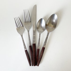 May include: A set of five stainless steel flatware pieces including two forks, one knife, and two spoons. Each utensil features a dark brown handle and a silver-toned metal top decorated with a delicate, repeating black filigree scroll pattern.