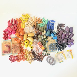 May include: A collection of colorful wooden beads in various shapes and sizes, including round, oval, and cylindrical. The beads are arranged in a rainbow of colors, with some packaged in vintage cards. A variety of craft supplies.