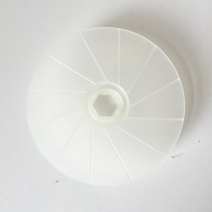 May include: A white plastic wheel with a central hexagonal hole and radiating spokes. The wheel is circular and appears to be made of a translucent material. The spokes divide the wheel into segments.