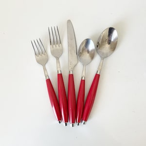 May include: A set of five pieces of stainless steel flatware with bright red handles. The set includes two forks, a knife, and two spoons. The utensils are arranged on a white surface.