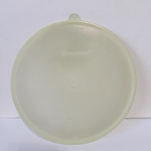May include: A round, translucent, pale yellow plastic Tupperware lid featuring a small tab at the top edge for easy removal. The surface is smooth with a subtle circular design in the center and the brand name embossed in the middle.