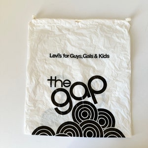 May include: White drawstring bag with the text "Levi's for Guys, Gals & Kids" and "the gap" in black. The bottom of the bag features a black circular design. The bag is crumpled and appears to be made of paper.