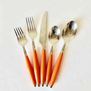 May include: A set of five stainless steel utensils with orange handles. The set includes two forks, a knife, and two spoons. The utensils are arranged on a white surface. The handles are a bright, solid orange color.