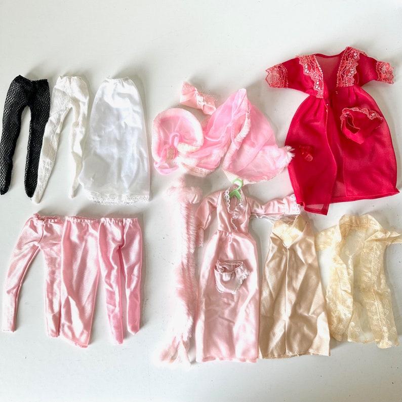 Op de afbeelding: Een verzameling roze, witte en rode poppenkleding, waaronder een slip, een nachtjapon, een badjas en een broek. De kleding is gemaakt van kant, satijn en tule.