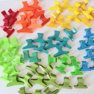 May include: A collection of plastic hair claw clips arranged by color, including red, orange, yellow, teal, light blue, and various shades of green. Each clip features a spring-loaded mechanism with interlocking teeth for securing hair sections.