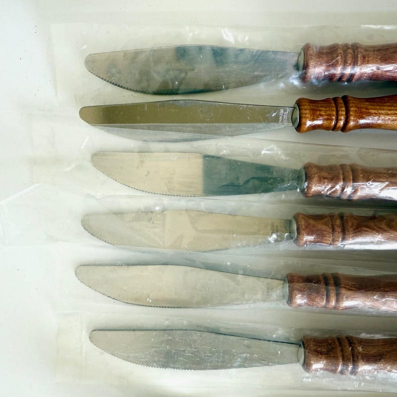 K&ouml;nnte beinhalten: Set mit sechs Buttermessern mit Edelstahlklingen und Holzgriffen. Die Klingen haben eine gezahnte Kante. Die Griffe sind mittelbraun und haben eine abgerundete Form. Die Messer sind einzeln in Klarsichtfolie verpackt.
