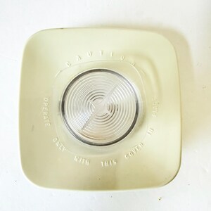 May include: A square, off-white plastic appliance cover featuring a circular, clear, ribbed center lens with a star-shaped detail. Embossed text reads: CAUTION OPERATE ONLY WITH THIS COVER IN PLACE. The cover has rounded corners and a smooth finish.