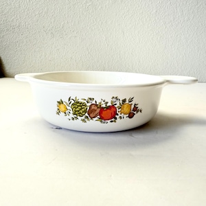 May include: White ceramic casserole dish with a colorful fruit and vegetable design. The design includes lemons, artichokes, tomatoes, and other produce. The dish has two handles and is suitable for baking and serving.