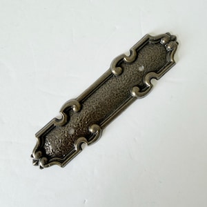 May include: An elongated, rectangular metal drawer pull backplate with a dark, antiqued bronze finish and a textured, hammered surface. The plate features decorative, curved scrollwork edges and two circular screw holes for mounting purposes.