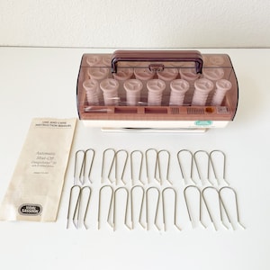 May include: A vintage Vidal Sassoon DesignSetter hair roller set. The set includes a brown and cream-colored heating unit with a clear lid, 20 pink rollers, and metal clips. An instruction manual is also included.
