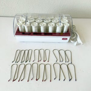 May include: A white and burgundy hair roller set with a clear plastic cover. The set includes multiple white rollers and metal hair pins with colored tips. The brand name "Vidal Sassoon" is visible on the front of the device.