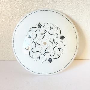 May include: A round, white glass ceiling light fixture with a decorative black floral pattern in the center. The edge has a dotted black border. The fixture is designed for overhead lighting.