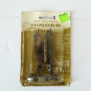 May include: A package of National Lock cabinet hinges with a hammered copper finish. The packaging is gold and white cardboard with clear plastic housing, featuring the text Quality Cabinet Accessories, HINGES, and the model number V363-100.