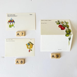 May include: Three vintage recipe cards with decorative illustrations. One card features a floral arrangement, another has a strawberry plant, and the third showcases a cuckoo clock. Scrabble tiles spell out "A, 7", "A, 8", and "A, 9".