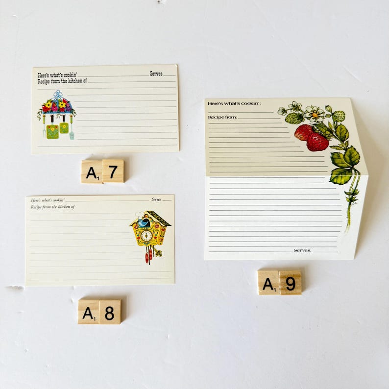 May include: Three vintage recipe cards with decorative illustrations. One card features a floral arrangement, another has a strawberry plant, and the third showcases a cuckoo clock. Scrabble tiles spell out "A, 7", "A, 8", and "A, 9".