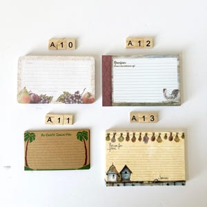 May include: Four recipe cards with decorative borders and lined pages. One card features fruit illustrations, another has a rooster, a third has palm trees, and the last has birdhouses and herbs. Scrabble tiles spell out A10, A11, A12, and A13.