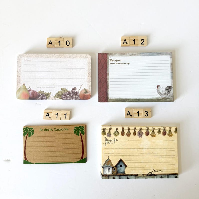May include: Four recipe cards with decorative borders and lined pages. One card features fruit illustrations, another has a rooster, a third has palm trees, and the last has birdhouses and herbs. Scrabble tiles spell out A10, A11, A12, and A13.