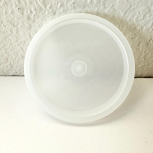 May include: A round, translucent white Tupperware lid. The lid has a slightly raised edge and a central circular indentation. The text "Tupperware" and "Made in U.S.A." are visible on the lid.