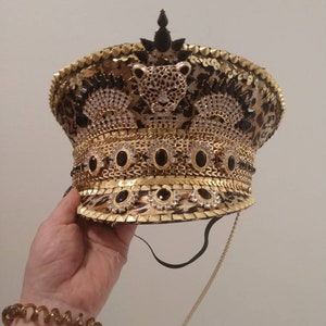 May include: A decorative, embellished hat with a leopard print pattern, gold sequins, and black and clear gemstones. The hat features a gold band and a leopard head emblem. A black chin strap and a gold cord are attached.