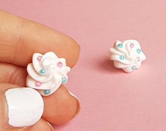 May include: A pair of white cupcake-shaped earrings with pink and blue sprinkles. The earrings are on a pink surface.