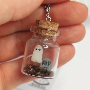 以下が含まれることがあります： コルク栓の付いたガラス瓶のネックレス。瓶の中には、黒い目の白い幽霊の置物と、「RIP」と書かれた小さな灰色の墓石が入っています。