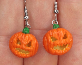 May include: A pair of orange pumpkin earrings with a green stem and a scary face. The earrings are on a silver hook.