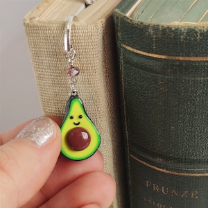 May include: A green avocado-shaped bookmark with a brown pit and a smiling face. The bookmark is attached to a silver chain with a pink crystal charm.
