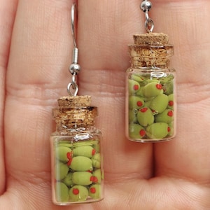 May include: A pair of dangle earrings featuring miniature glass jars filled with green olives. The jars have cork stoppers and are suspended from silver hooks.