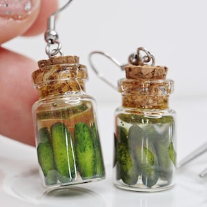 Pode incluir: Um par de brincos pendentes com miniaturas de frascos de vidro cheios de fatias de picles verdes. Os frascos têm tampas de cortiça e são presos a ganchos de brincos de prata.