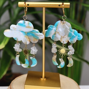 Puede incluir: Pendientes de tono dorado con un diseño de nube de mármol azul y blanco y colgantes que incluyen un sol, una luna y piedras de cristal.