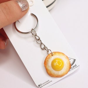 Op de afbeelding: Een sleutelhanger in de vorm van een gebakken ei gemaakt van polymer clay. Het ei is geel met een wit eigeel en een bruine korst. De sleutelhanger is bevestigd aan een zilveren metalen ring en ketting.