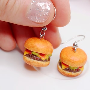 Op de afbeelding: Miniatuur hamburger oorbellen met zilverkleurige haken. Elke oorbel heeft een gedetailleerde hamburger met een sesambroodje, kaas, sla, tomaat en een hamburger patty. De oorbellen zijn een leuk en uniek accessoire.