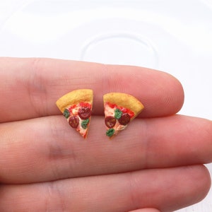 May include: A pair of miniature pizza slice earrings. Each earring features a golden-brown crust, red tomato sauce, pepperoni, and green basil. The earrings are small and detailed, perfect for a food-themed jewelry collection.