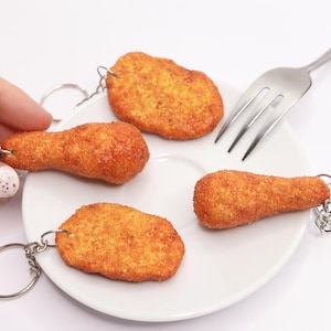 Könnte beinhalten: Vier neuartige Lebensmittel-Schlüsselanhänger, zwei Hähnchenschenkel und zwei Chicken Nuggets, liegen auf einem weißen Teller. Eine silberne Gabel liegt auf dem Teller. Die Schlüsselanhänger sind an silbernen Schlüsselringen befestigt.