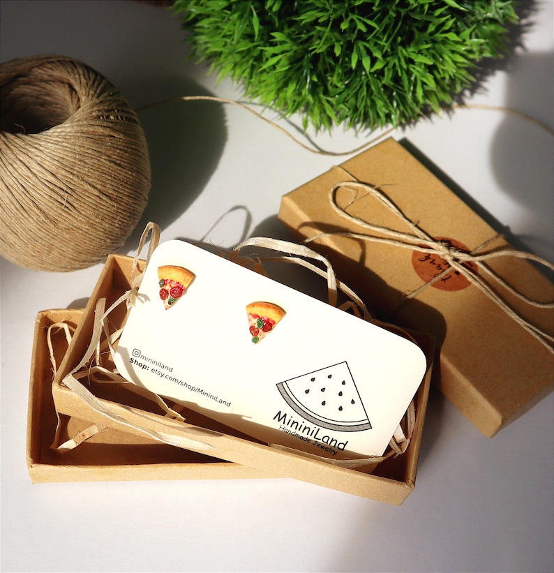 May include: Pizza slice earrings on a white card inside a small brown box. The earrings are shaped like pizza slices with red and green toppings. The card has the shop name and a watermelon illustration. A brown twine is wrapped around the box.