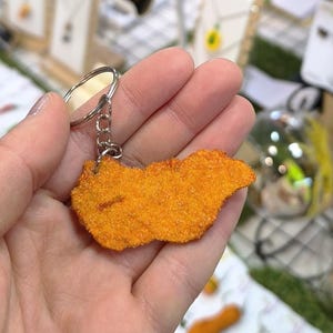 Peut inclure: Un porte-clés fantaisie avec un charm en forme de nugget de poulet orange. Le charm est attaché à un porte-clés et une chaîne argentés. Le nugget a une surface texturée, imitant un aliment frit. Un accessoire amusant et unique.