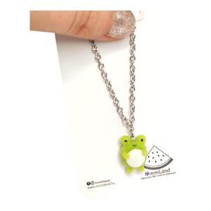 May include: A silver chain necklace with a green frog charm. The frog is holding a white circle. The necklace is on a white background with the text "@mininiiland" and "www.mininiiland.hu" at the bottom. The text "MininiLand" is at the bottom right corner of the image.
