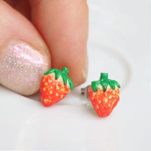 Pode incluir: Um par de brincos de morango. Os brincos são vermelhos com pequenas sementes amarelas e topos verdes. Os brincos estão em uma superfície branca.