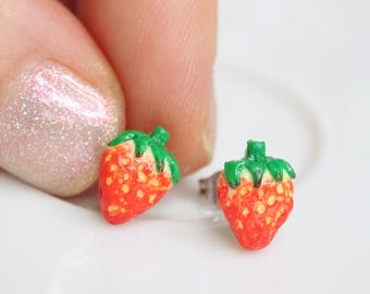 May include: A pair of strawberry-shaped stud earrings. The earrings are red with small yellow seeds and green tops. The earrings are on a white surface.