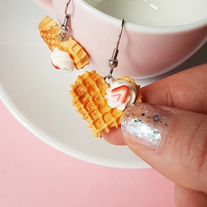 Könnte beinhalten: Ein Paar herzförmige Waffel-Ohrringe mit einem weißen und roten Erdbeer-Topping. Die Ohrringe sind aus Polymer Clay gefertigt und haben silberne Haken.