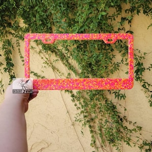 May include: Pink and orange glitter license plate frame. The frame is made of plastic and has a shiny finish.